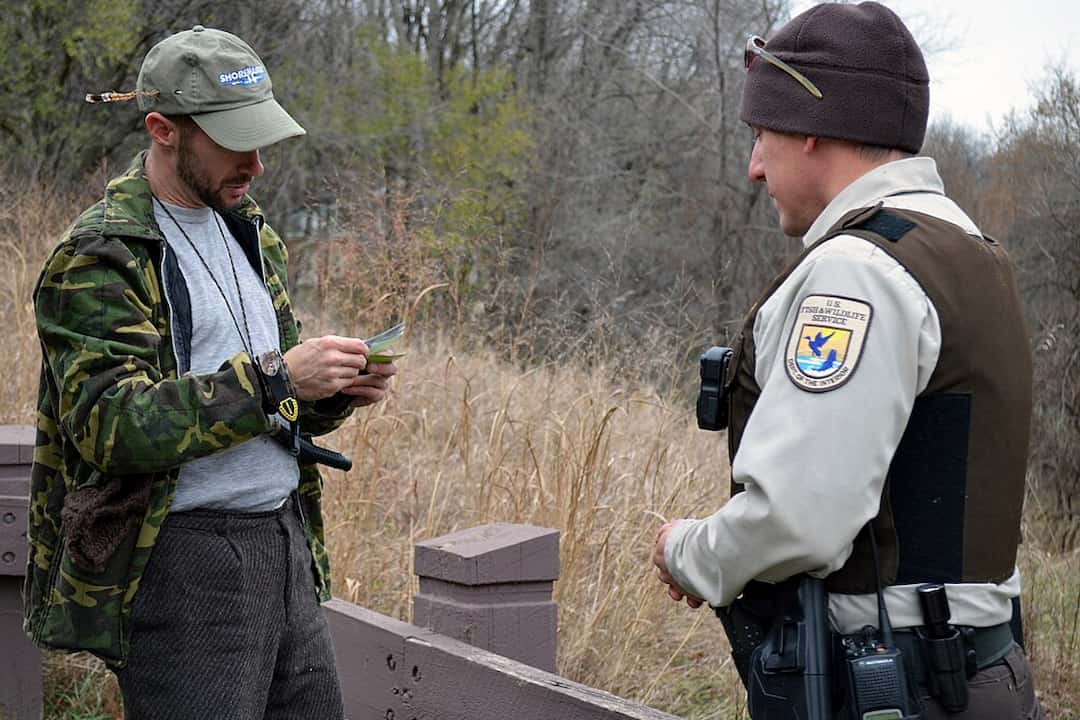 U.S. Fish and Wildlife officer checking a hunter's license outdoors in a grassy area, with both individuals dressed in cold-weather gear
