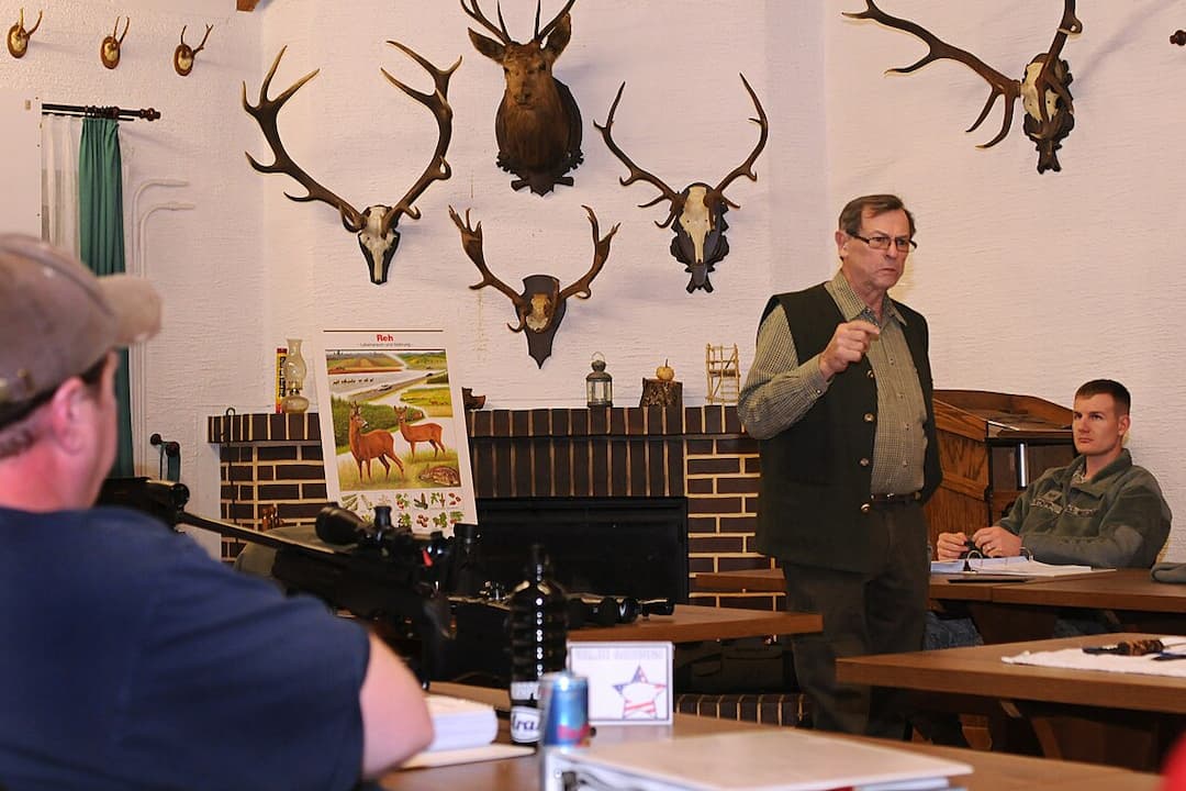  Indoor hunting education session with mounted deer antlers on the wall, an older man standing and instructing, several seated students taking notes, and a poster showing deer anatomy