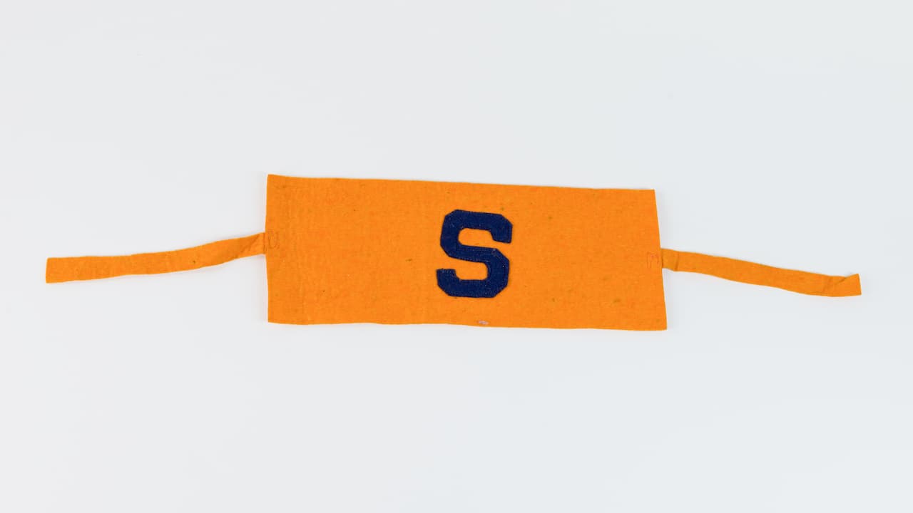 Orange fabric armband, dark blue letter “S” in center, rectangular panel with two tie straps, laid flat on white background