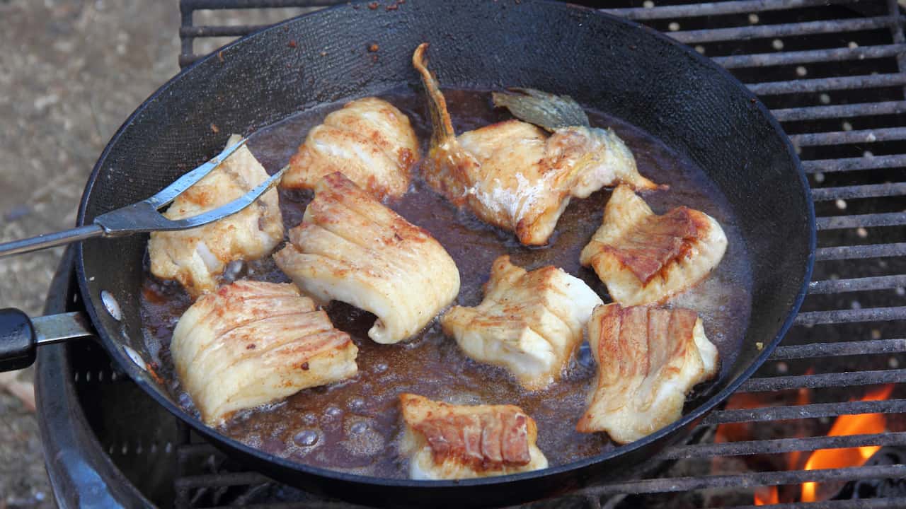 Chunks of white fish frying in a cast-iron skillet over an open flame, surrounded by bubbling sauce