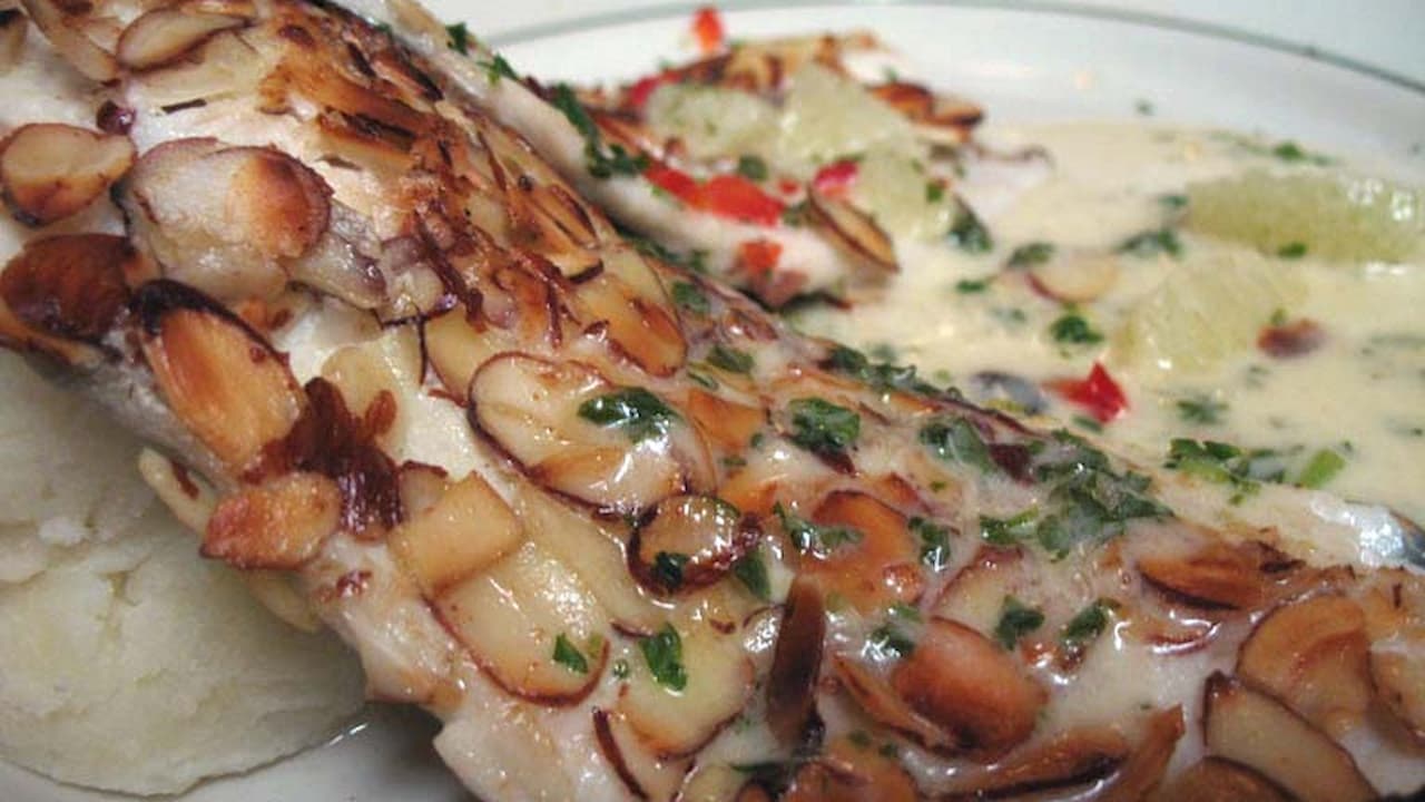 Close-up of trout almondine fillet topped with toasted almond slices and herbs, served in a creamy sauce with lemon wedges and vegetables