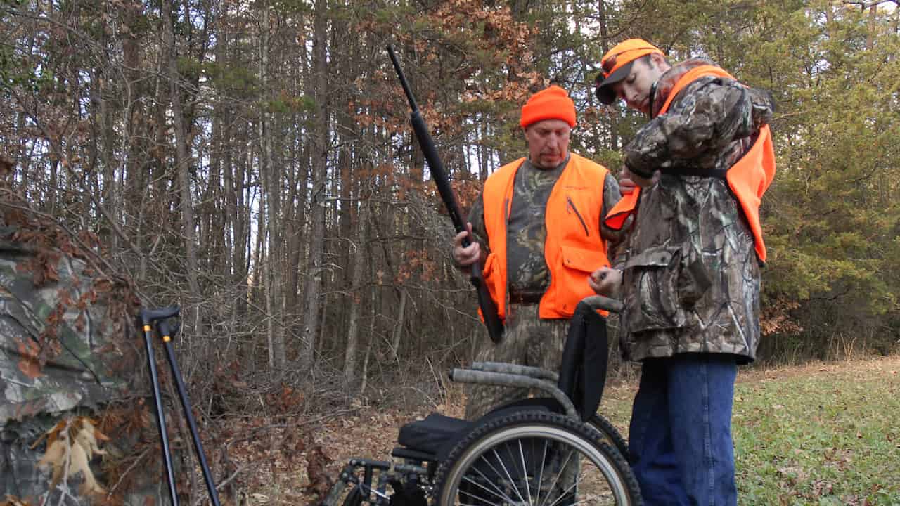 Two hunters in orange safety vests and camouflage clothing, one holding a rifle, standing beside a wheelchair and a pair of crutches, forest background with fallen leaves and autumn trees