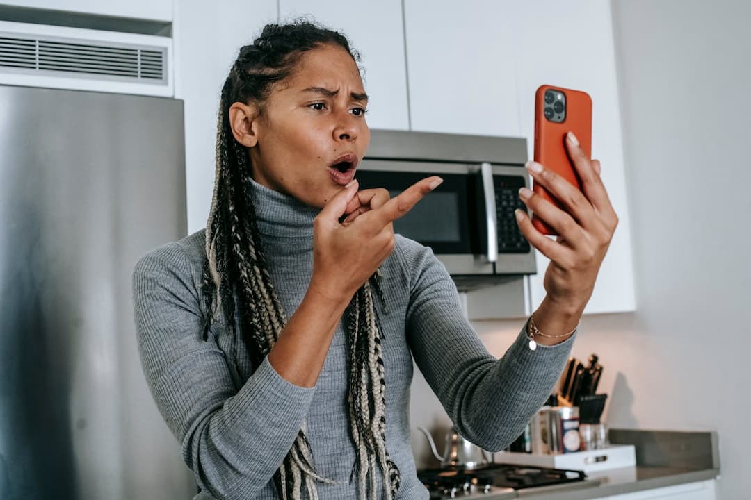 woman standing in a kitchen, holding a smartphone with an orange case, looking at it with a shocked expression and gesturing angrily with her hand