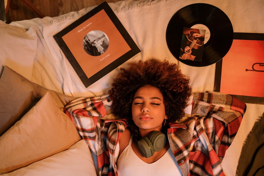 young woman with curly hair relaxing on a bed with eyes closed, wearing a plaid shirt and headphones around her neck, surrounded by vinyl records and pillows