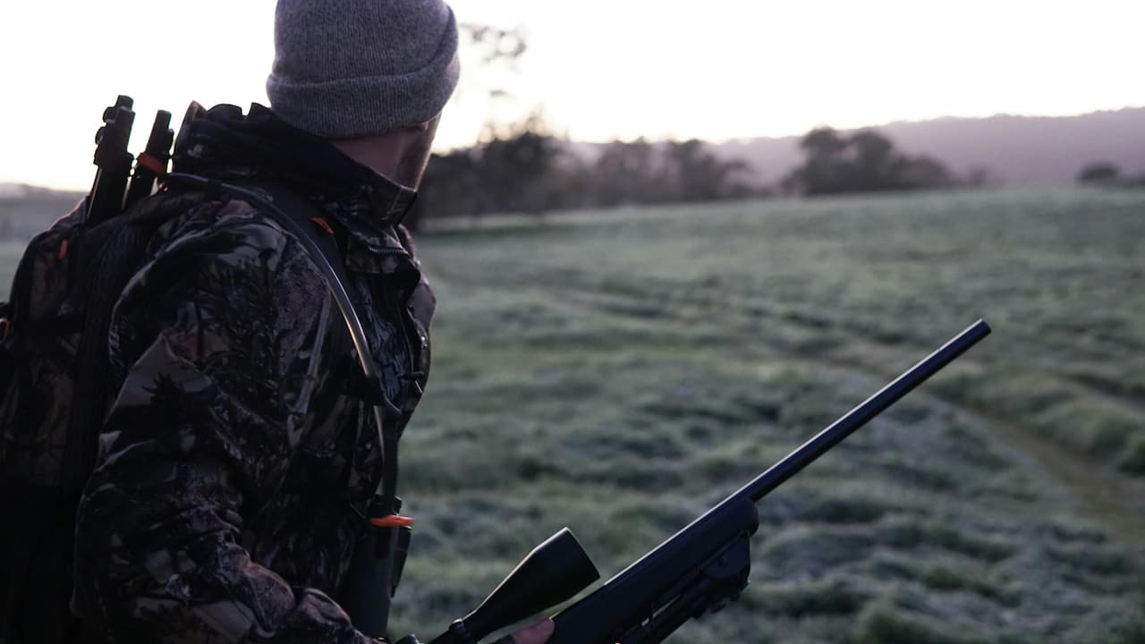 Person in camouflage jacket and gray beanie holding rifle, carrying backpack with arrows, standing in frosty field at dawn, looking toward distant trees