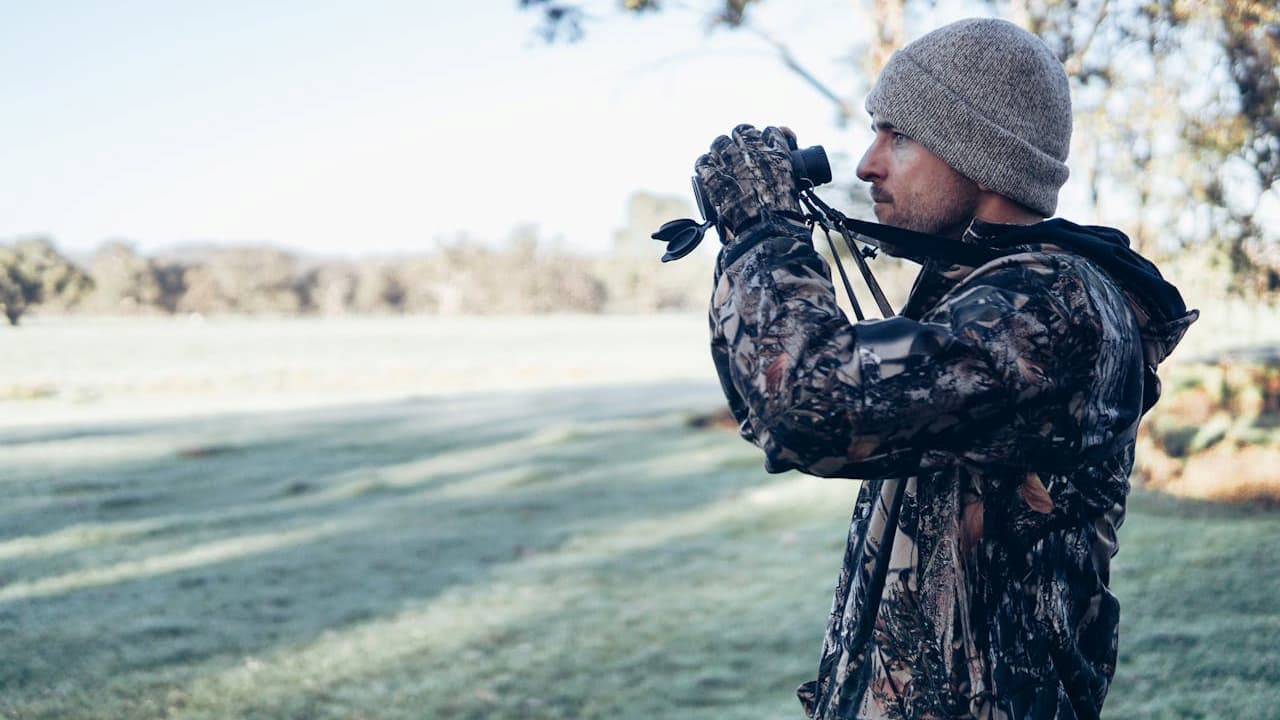 Man in camouflage jacket and knit hat using binoculars in a grassy outdoor setting
