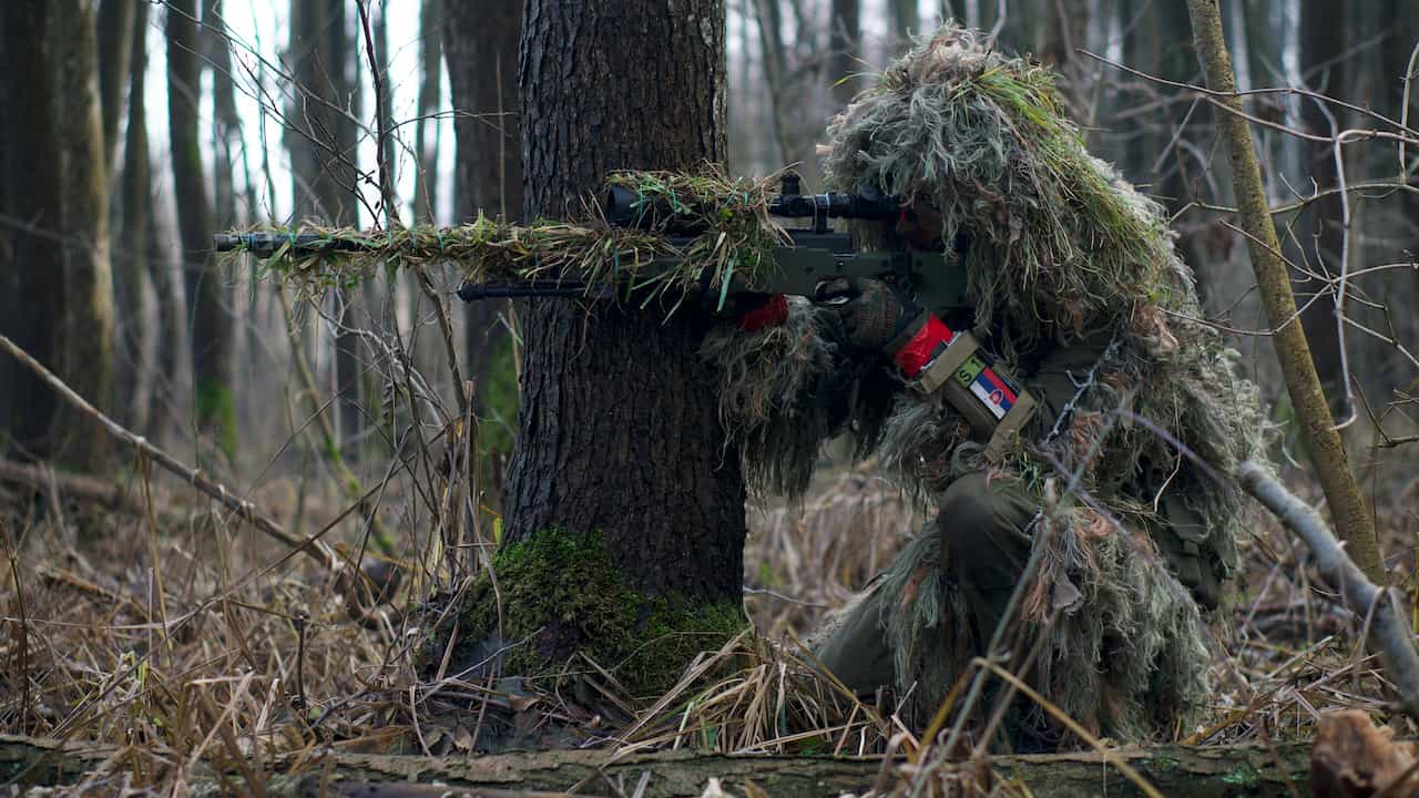 Camouflaged sniper in a ghillie suit aiming rifle in forest