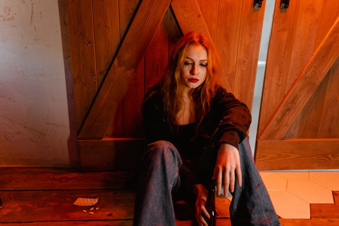 woman with red hair sitting on the floor in dim lighting, looking distressed, holding a beer bottle, with scattered pills and an empty blister pack nearby