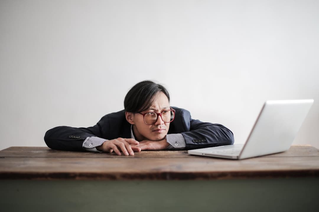 man in glasses leaning on a wooden desk, staring at a laptop with a frustrated or bored expression