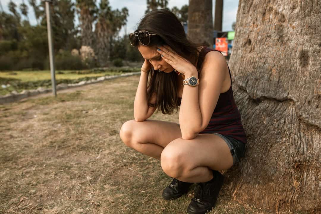woman squatting beside a tree with her hands on her head, appearing stressed or overwhelmed