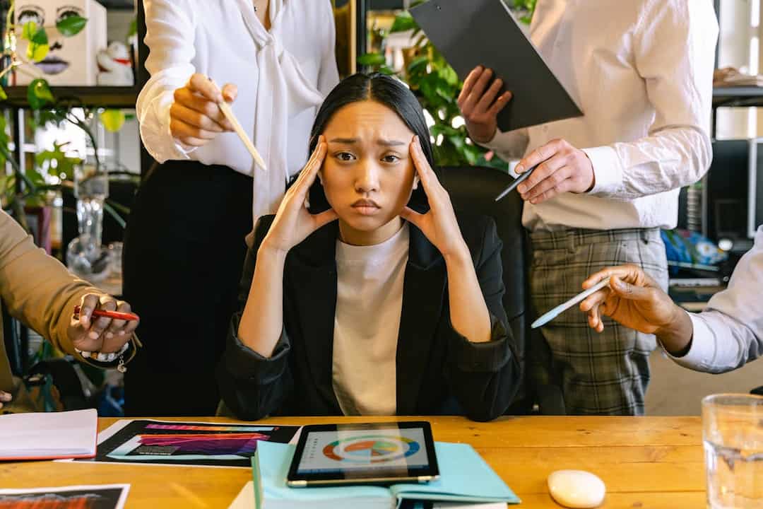 overwhelmed woman in an office surrounded by coworkers pointing pens and documents at her, holding her temples with a frustrated expression, sitting at a cluttered desk with a tablet and charts
