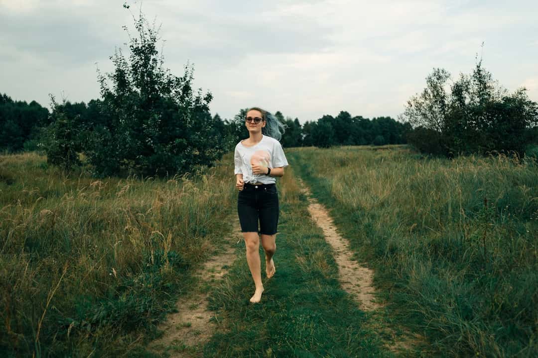 barefoot woman jogging on a narrow dirt path through a grassy field, wearing sunglasses, a white t-shirt, and black shorts