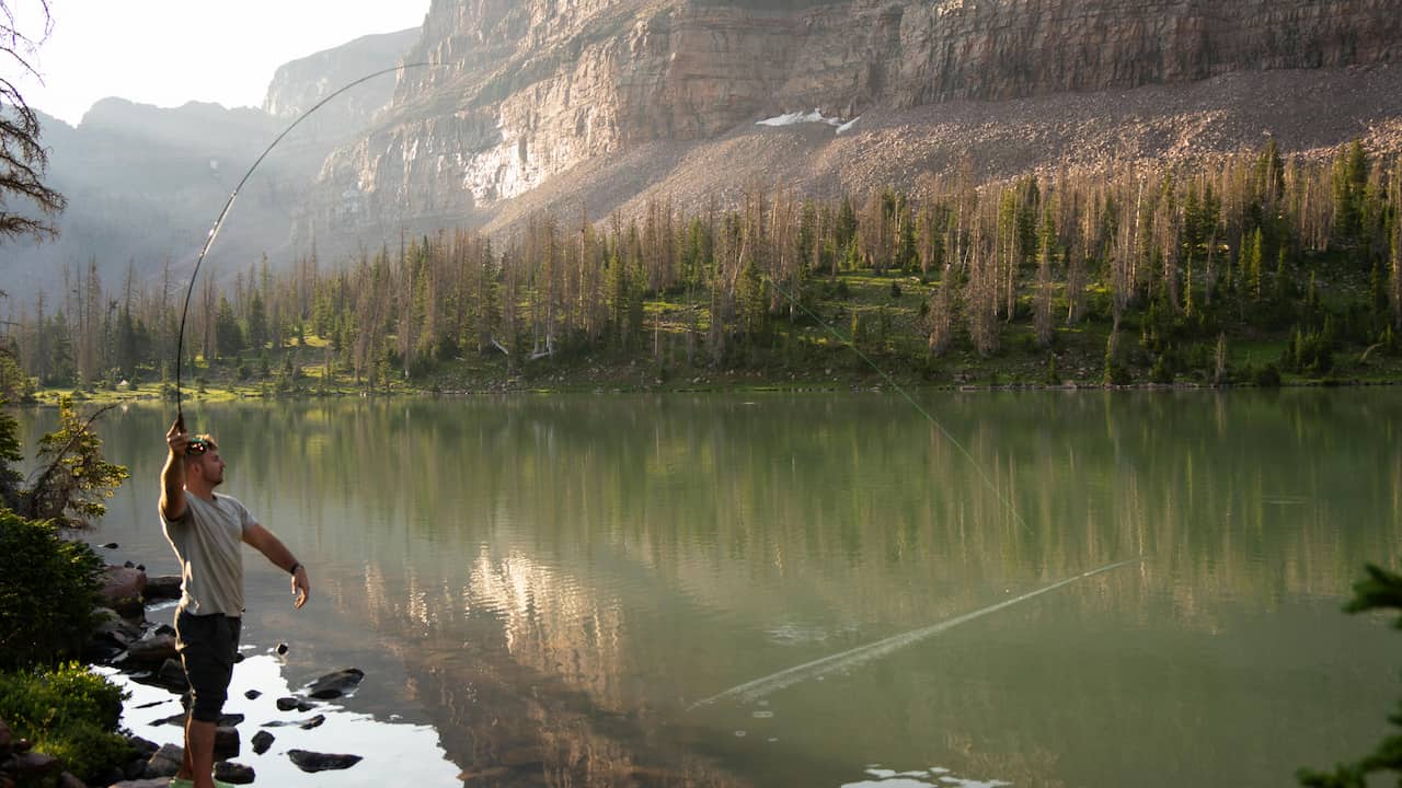 Man fly fishing in mountain lake, casting rod with curved line, standing on rocky shore, calm green water, forested slope and rocky cliffs in background