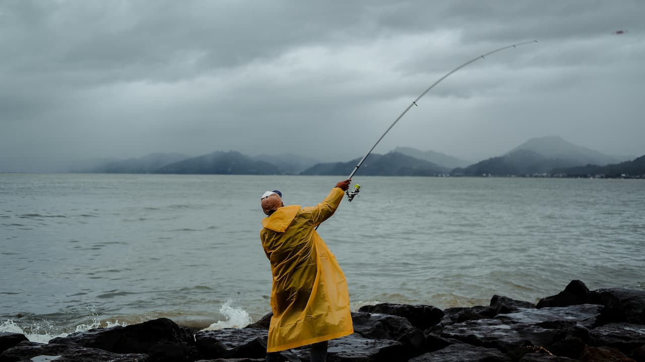 Person fishing from rocky shoreline, wearing yellow raincoat and cap, casting rod into ocean, cloudy sky, misty mountains in background