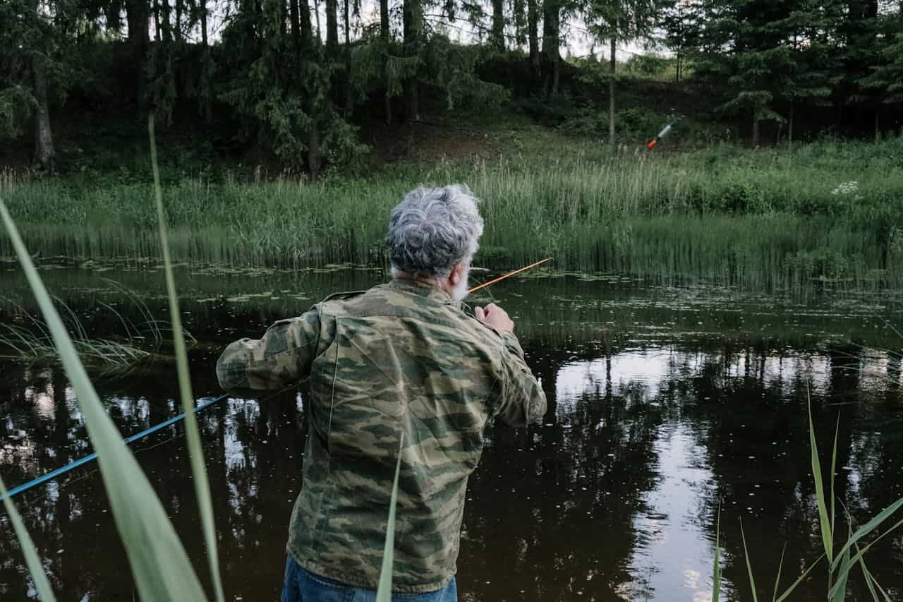 Older man fishing by pond, wearing camouflage jacket, casting line into still water, surrounded by reeds and dense greenery, forest in background