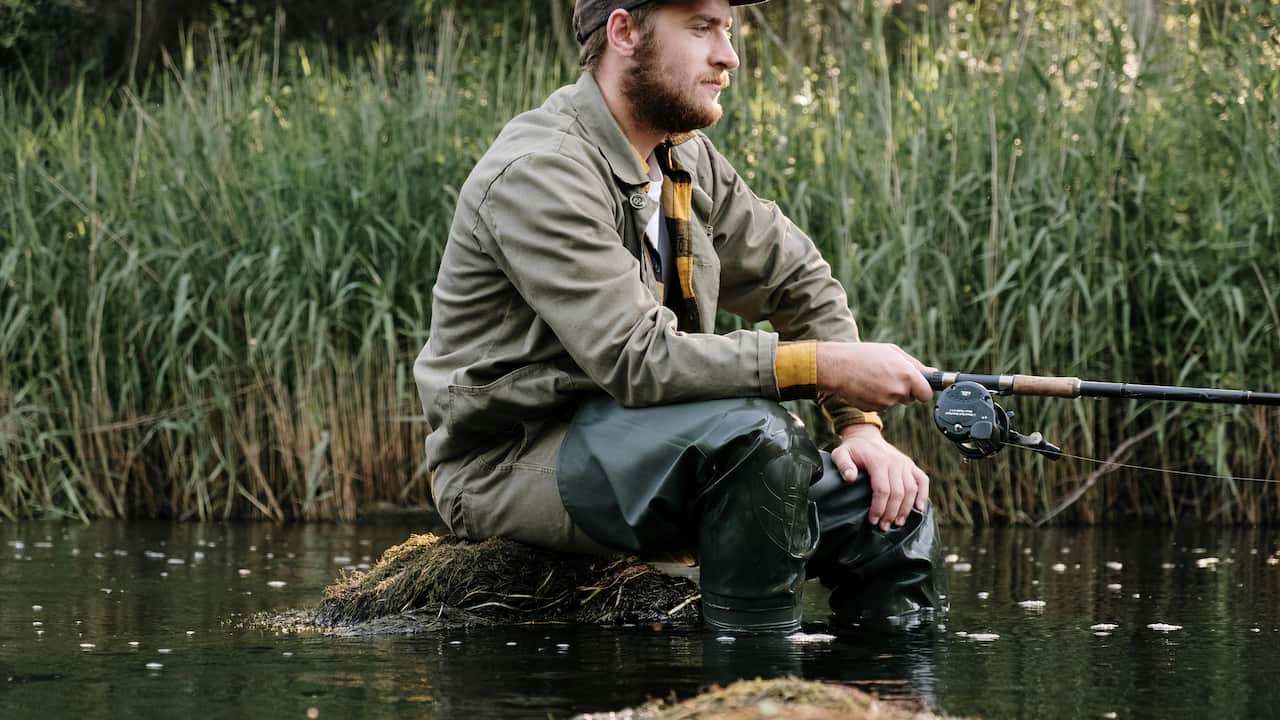 Man fishing while kneeling on mossy rock, holding rod with baitcasting reel, wearing waders, jacket, and cap, tall grass in background, calm water surface