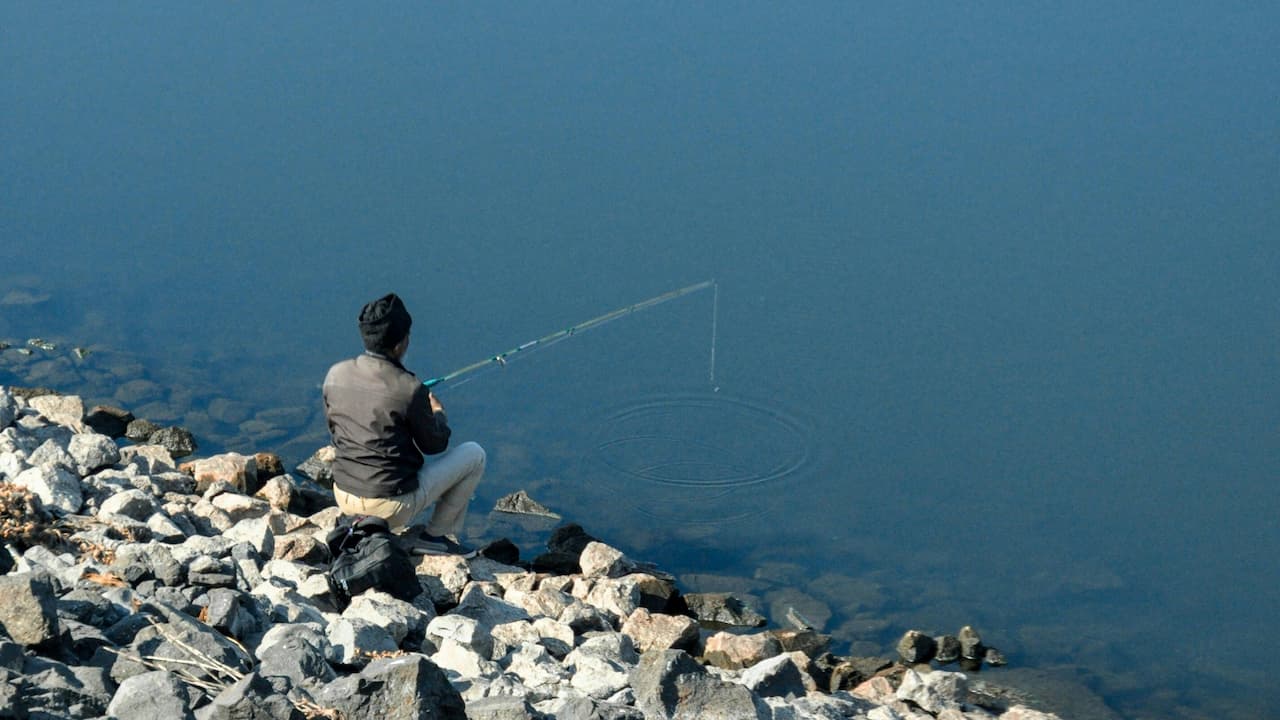 Person fishing alone by calm lake, seated on rocky shore, wearing dark jacket and beanie, fishing rod extended over still water with visible ripples
