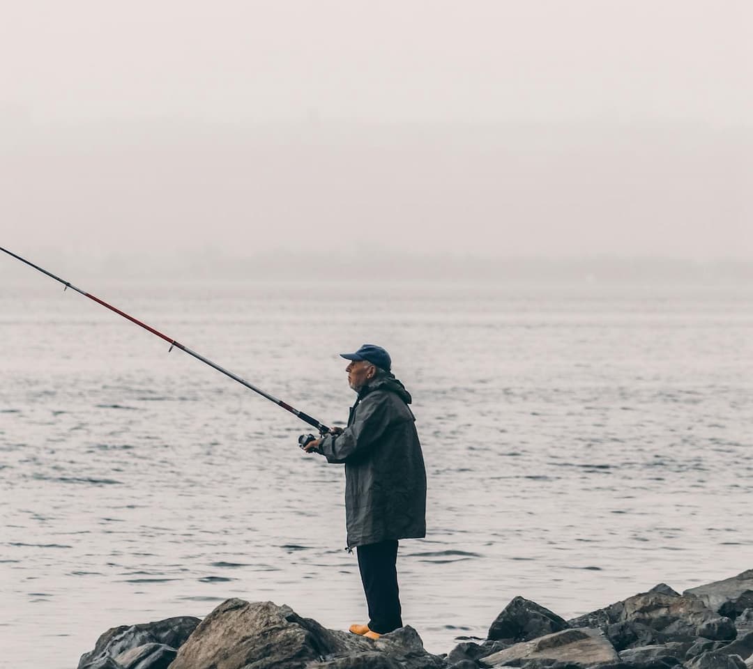 Man in a raincoat and cap standing on coastal rocks, fishing with a long rod over a calm, misty body of water