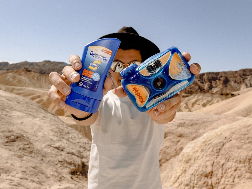 Person outdoors holding sunscreen and camera, wearing a wide-brim hat for sun protection