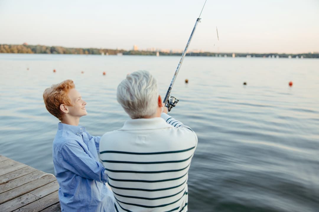 Older man casting a fishing line from a dock while a smiling red-haired youth looks on beside him