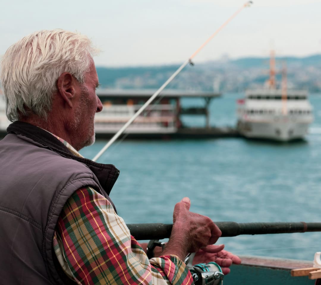Elderly man with white hair and a plaid shirt reeling in a fishing rod while standing near a dock, with ferry boats and a cityscape in the background
