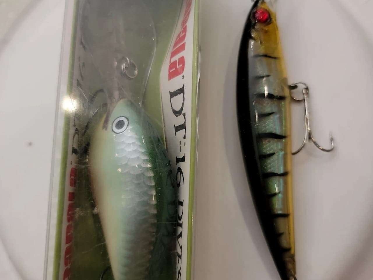 Two fishing lures on a white surface; one in packaging with a silver-green body and the other with a black-striped translucent body