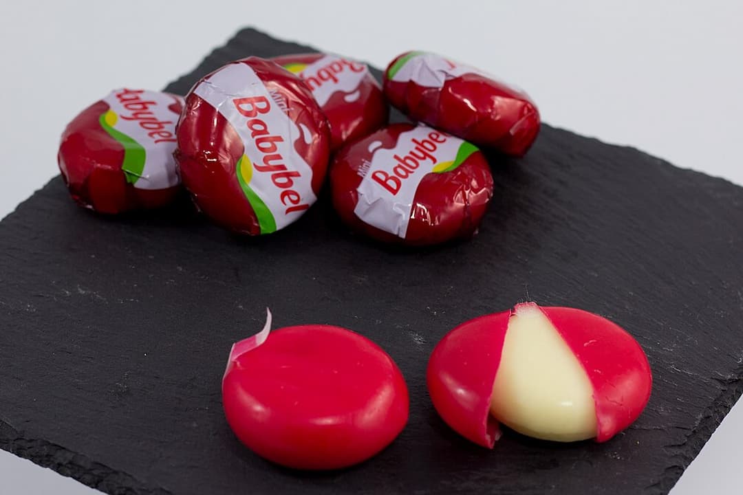 Several Babybel mini cheeses in red wax wrappers on a dark slate surface, with one cheese partially unwrapped to reveal creamy white cheese inside