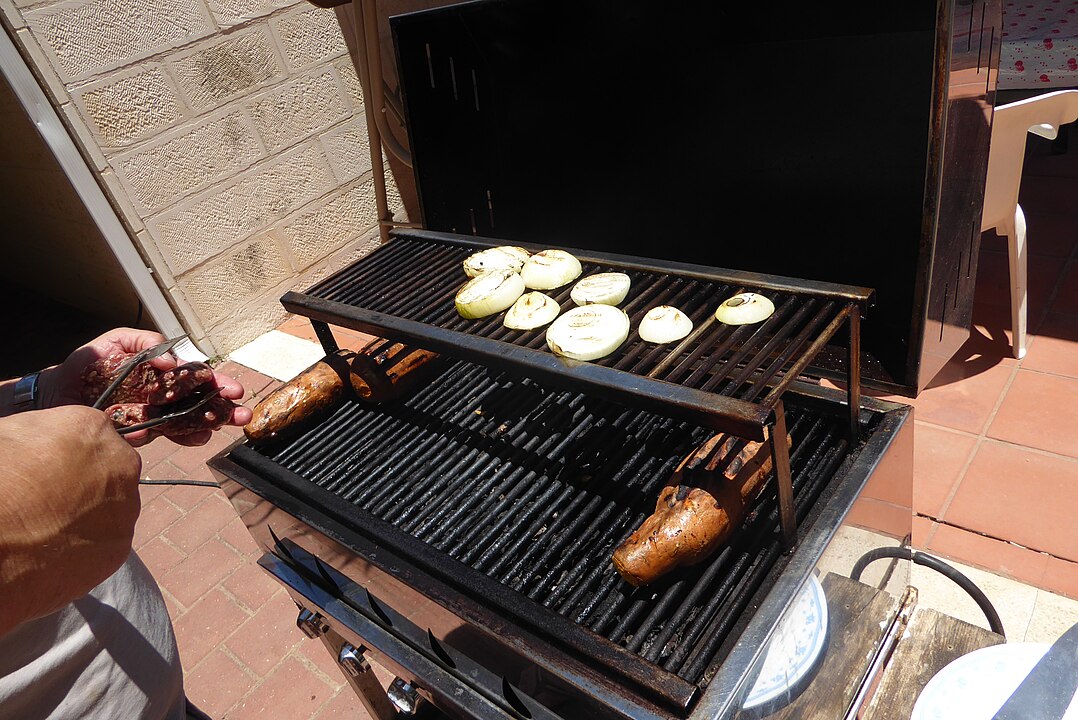 Grill with sausages on the lower rack and sliced onions cooking on the upper rack, person using tongs to handle meat beside the barbecue