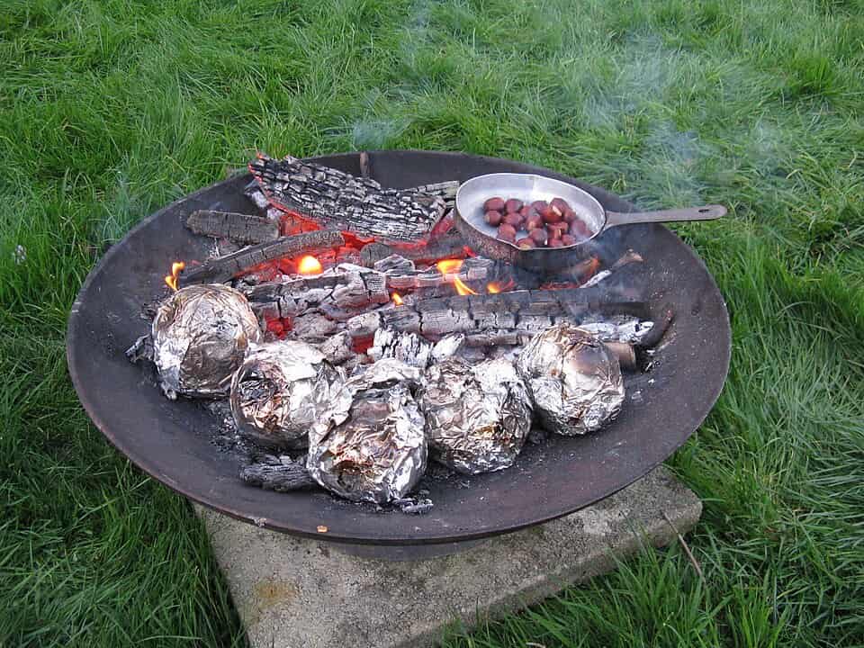 Chestnuts in a metal pan and foil-wrapped potatoes roasting on an open fire pit with glowing embers