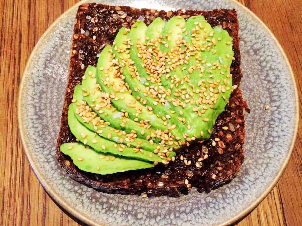 Slice of dark whole grain toast topped with thinly sliced avocado and sprinkled with sesame seeds, served on a ceramic plate