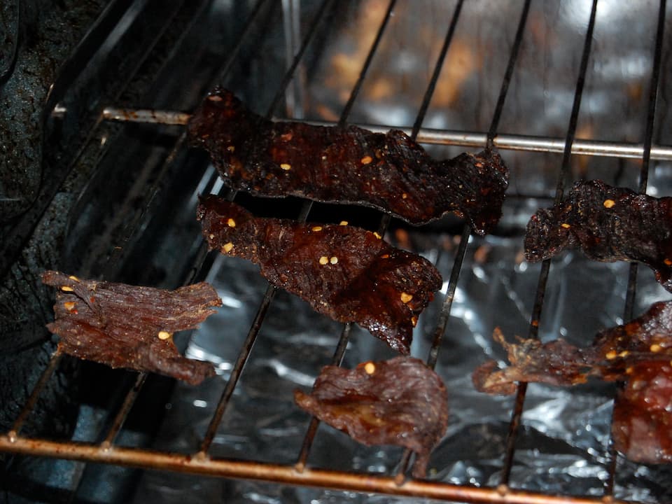 Beef jerky strips drying on an oven rack, dark in color with visible chili flakes, above foil-lined tray