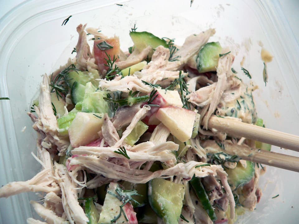 Shredded chicken salad with diced apples, cucumbers, and herbs in a plastic container
