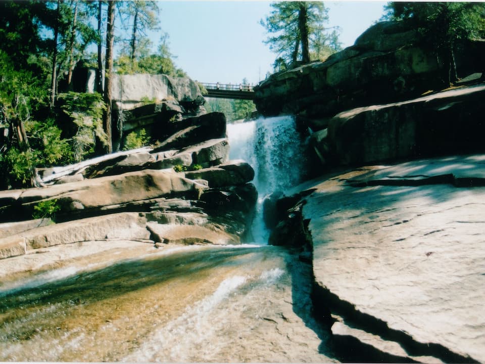 Waterfall cascading beneath a wooden footbridge surrounded by large granite rocks and tall pine trees in Yosemite National Park