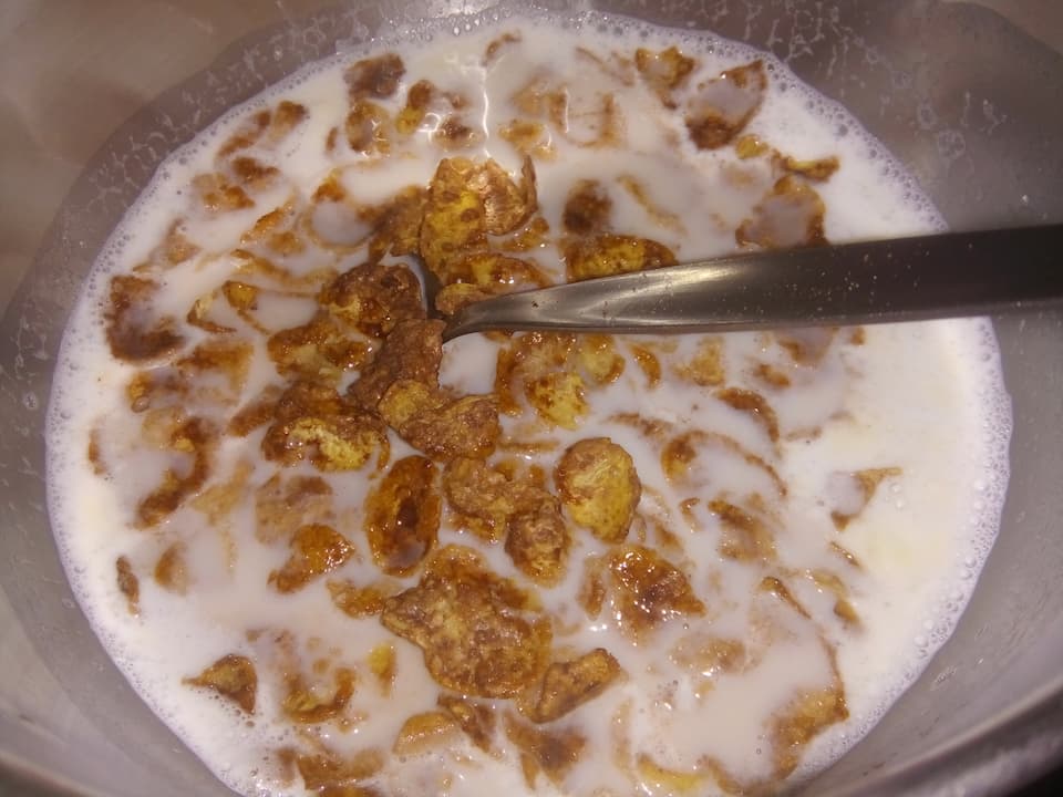 Bowl of brown cereal flakes with powdered milk, partially soaked in milk, with a metal spoon resting inside