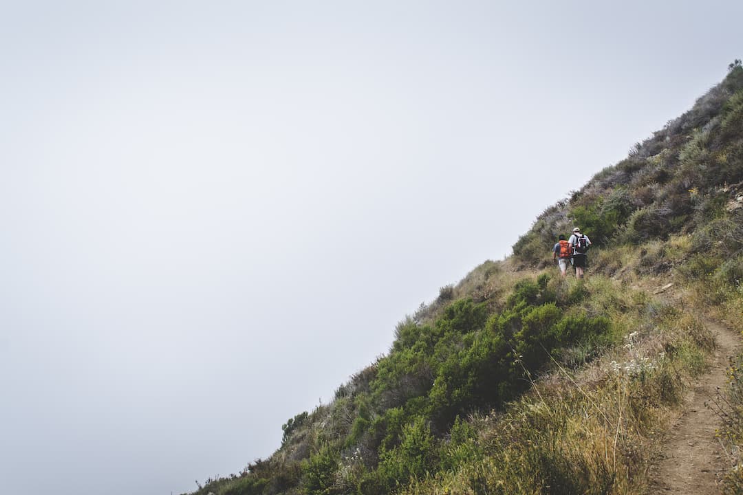 Two hikers with backpacks walking uphill on a narrow dirt trail along a steep, shrub-covered slope under a cloudy sky