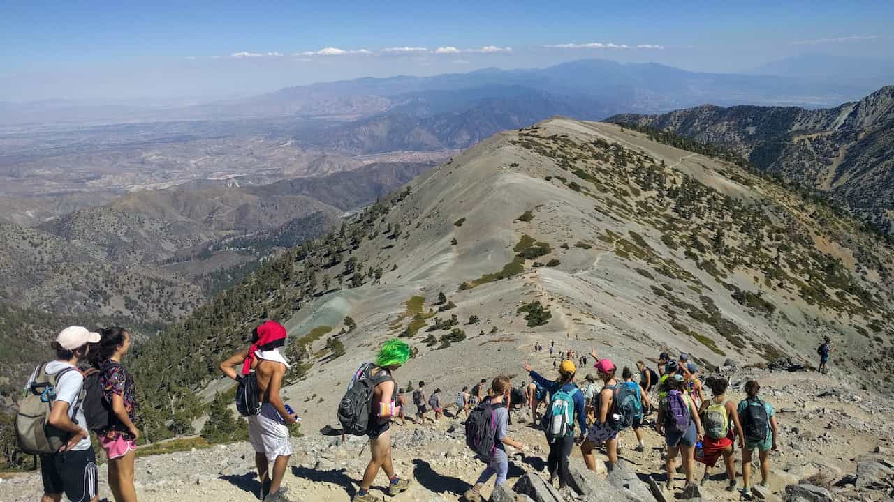 A large group of hikers wearing backpacks walks in single file along a narrow mountain ridge trail with a panoramic view of valleys and peaks under a clear sky