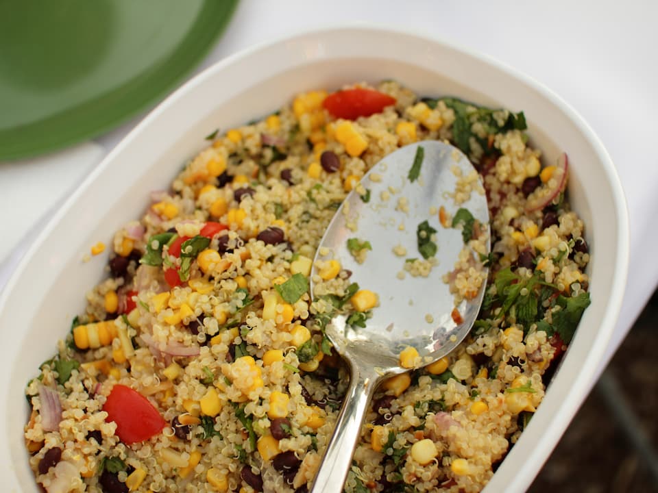 Quinoa salad in a white dish, mixed with corn, black beans, chopped tomatoes, onions, and fresh herbs, with a serving spoon resting on top