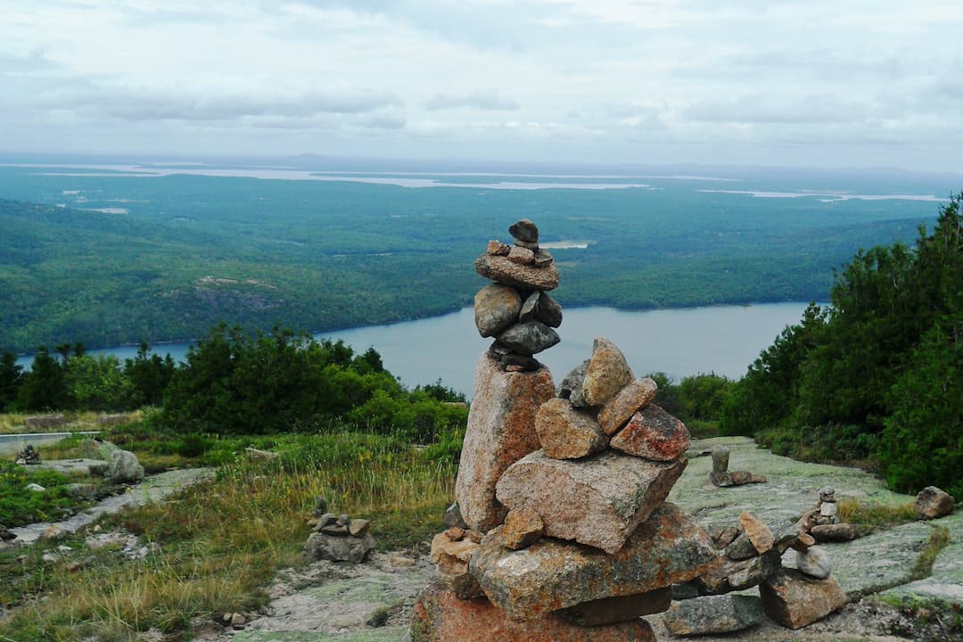 Stacked rocks forming cairns on a rocky hilltop, overlooking a lush forested landscape with a lake and distant hills under a cloudy sky