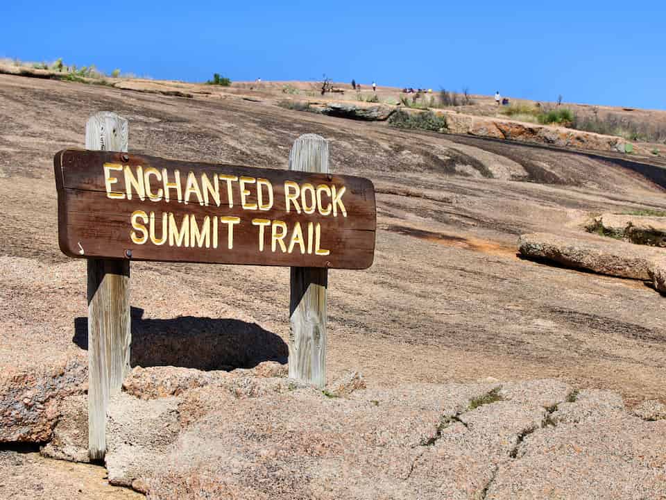 Wooden sign for Enchanted Rock Summit Trail on a large granite rock surface under a clear blue sky