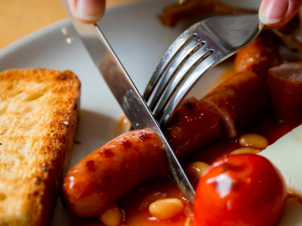 Close-up of sausage being sliced with a fork and knife, served with baked beans, cherry tomato, toast, and melted cheese on a white plate