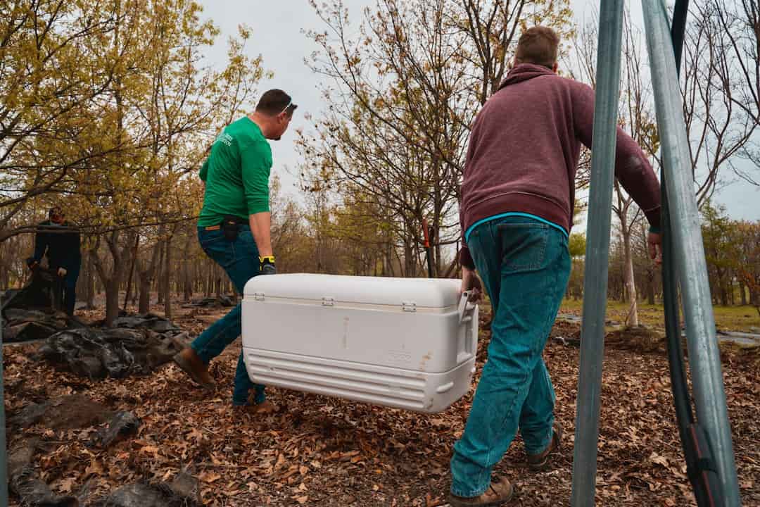Two men carrying a large white cooler through a wooded area with dry leaves, wearing jeans and jackets, one in green and the other in maroon, during a camping or outdoor event