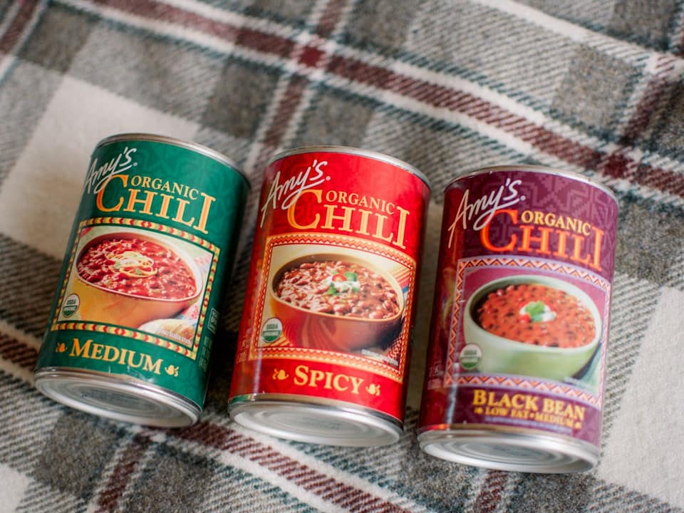 Three cans of Amy’s Organic Chili placed on a plaid fabric surface, labeled Medium, Spicy, and Black Bean Low Fat Medium, each featuring a bowl of chili on the front