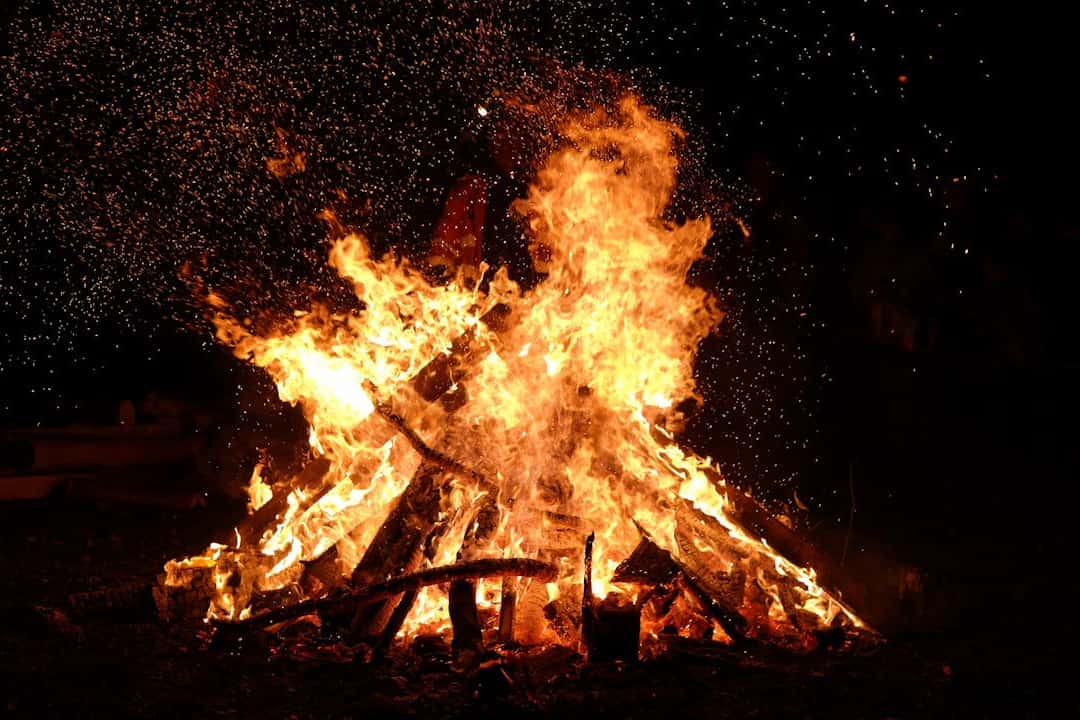 Large campfire burning at night, tall orange flames rising from stacked logs, glowing embers scattering in the dark, faint silhouettes of people in the background