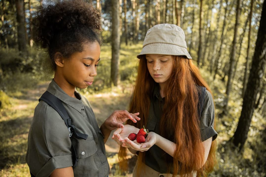 Two girls in outdoor clothing examining strawberries in a forest, one holding the berries in her palm, the other reaching to touch them, surrounded by tall trees and soft daylight