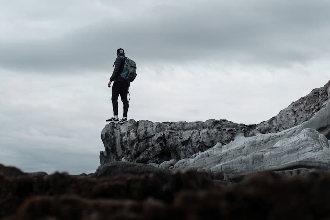 Man wearing a backpack standing on a rugged rock formation, looking into the distance under a cloudy sky