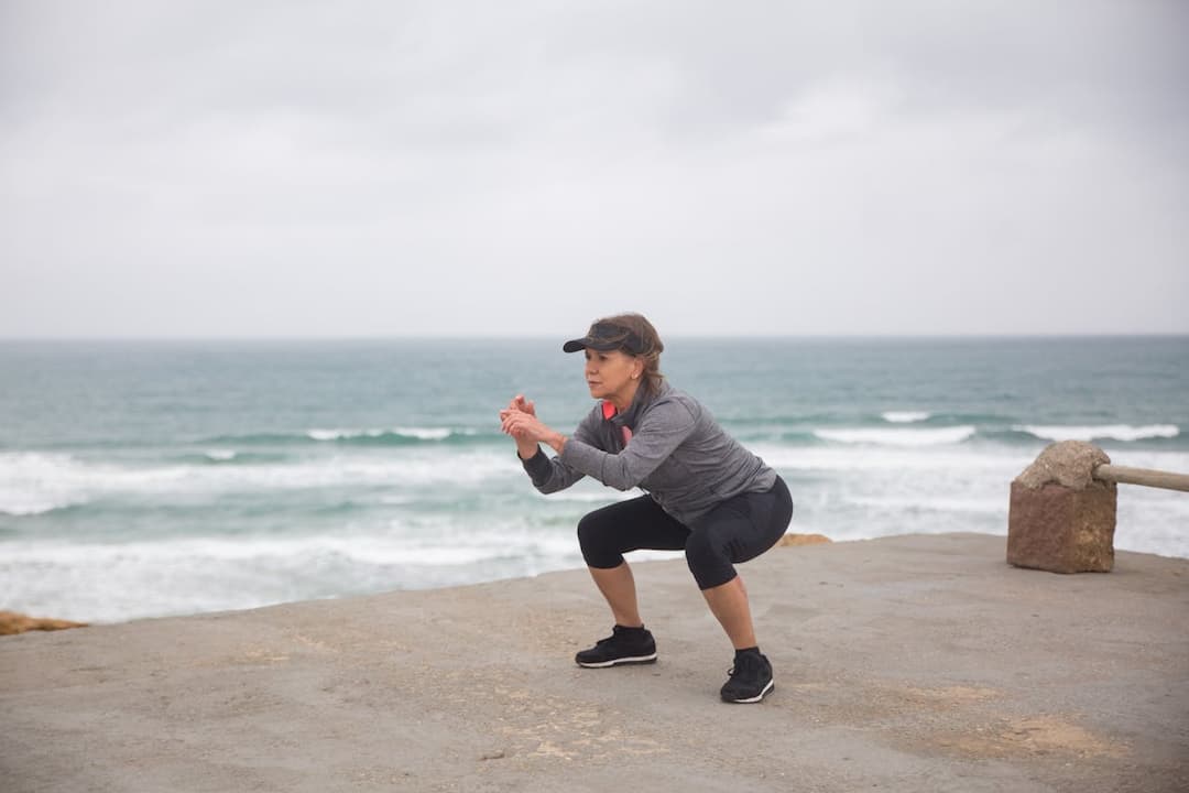 Woman wearing athletic gear performing a squat on a concrete surface near the ocean, with waves in the background under an overcast sky