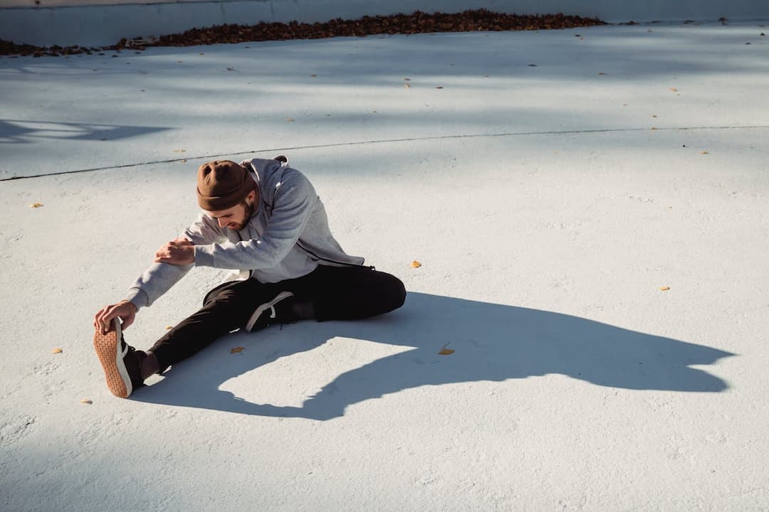 Man in a beanie and hoodie sitting on the ground stretching his leg forward, reaching for his foot on a smooth outdoor surface with scattered leaves
