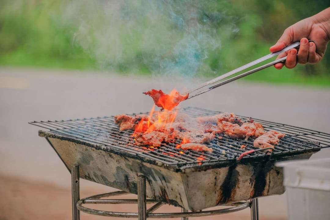 Outdoor barbecue grill with flames and smoke, person using metal tongs to flip grilled meat