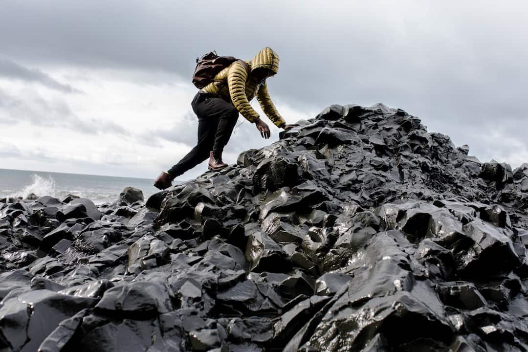 Person in a yellow hooded jacket and brown backpack climbing shiny black volcanic rocks near the ocean under a cloudy sky