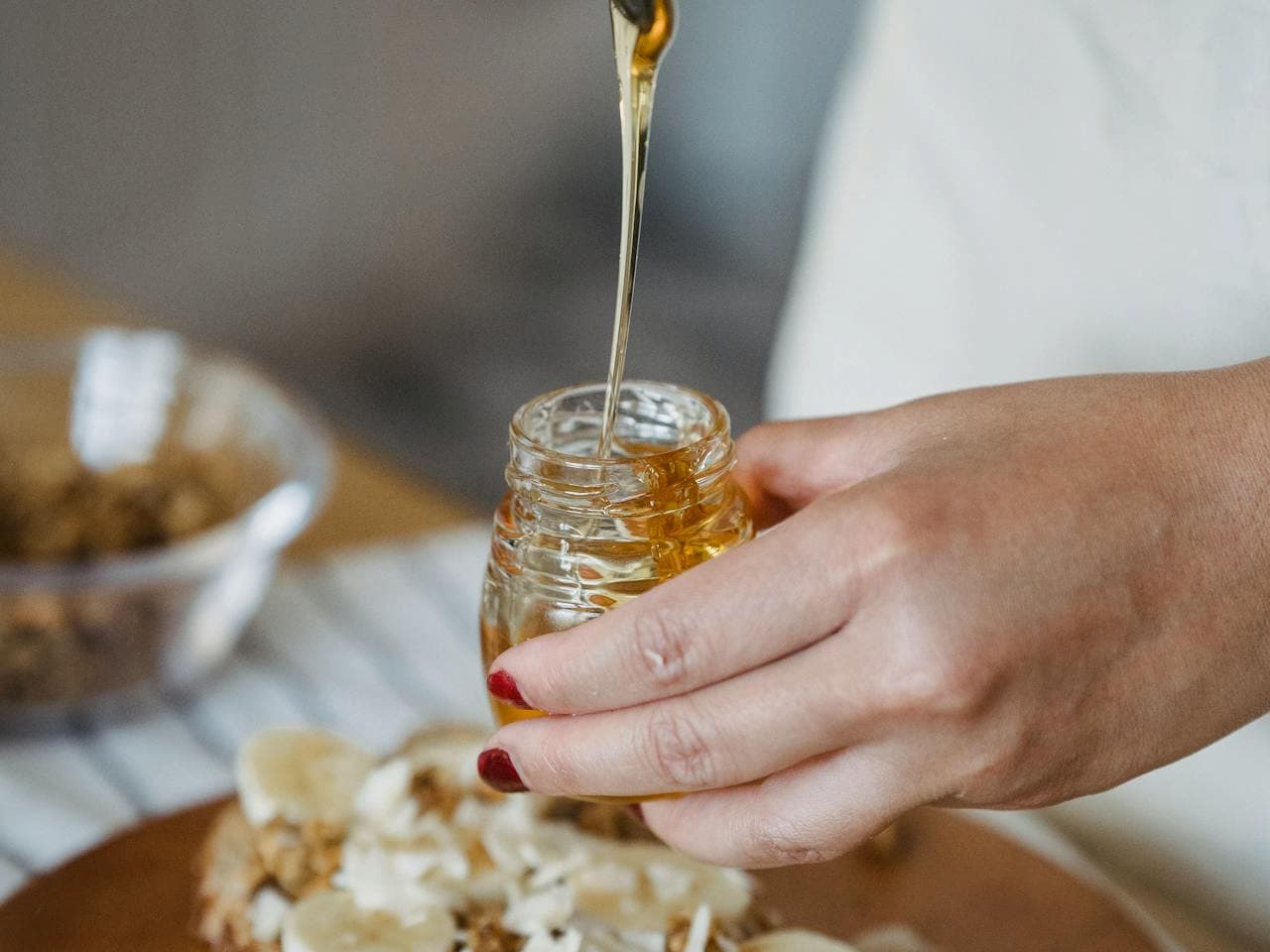 Close-up of a hand holding a small glass jar of honey, with honey being drizzled onto a sandwich topped with banana slices and shredded coconut